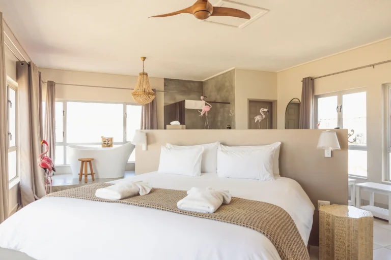 Luxury open-plan bedroom suite featuring a freestanding white bathtub, neutral beige decor, a beaded chandelier, and whimsical pink flamingo accents.