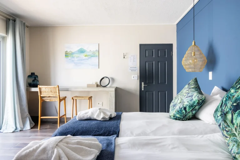 Modern guest room interior featuring a blue accent wall, tropical leaf print pillows, and minimalist wicker furniture.